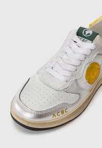 Zapatilla con parte superior de cuero plateado, cordones blancos, logo circular amarillo y suela de goma negra texturizada. El diseño incluye detalles perforados.