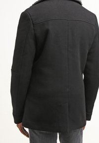 Svart ullrock med en texturerad krage, skräddarsydd passform och sidofickor. Har en ren design med subtila sömmar och rak rygg.