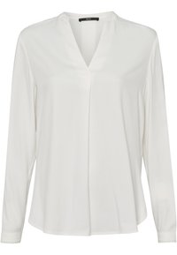 Blouse blanche à manches longues avec col en V, coupe ample et pli discret à l'avant, étiquetée "zero" au col.