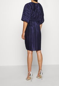 Plisserad lila klänning med korta ärmar, åtsittande midja och knälång nederkant. Har en slät textur och subtil lyster. Matchas med silverfärgade klackar.