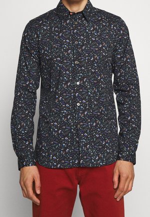 Muž v dlouhém černém tričku s drobným květinovým vzorem a jasně červených kalhotách stojí s rukama volně podél těla.