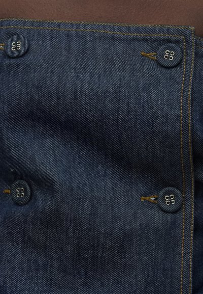 Σκούρο μπλε πάνελ από τζιν ύφασμα με τέσσερα κουμπιά καλυμμένα με ύφασμα που ράβονται στις γωνίες, στερεωμένα με καφέ κλωστή.