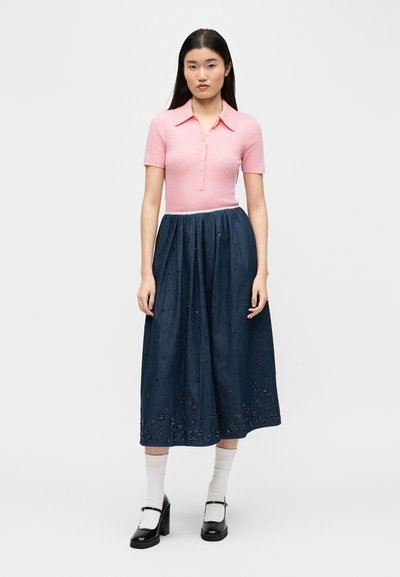 Γυναίκα που στέκεται, φορώντας ροζ κοντομάνικο πουκάμισο με κουμπιά, μεσάτο φόρεμα σε μπλε ναυτικό χρώμα με κεντήματα, λευκές κάλτσες και μαύρα παπούτσια Mary Jane.