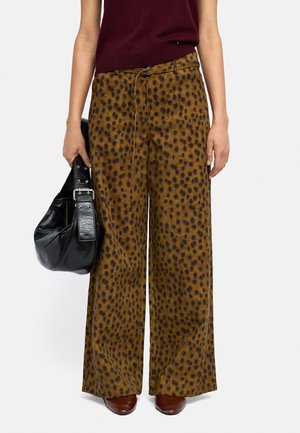 Osoba w brązowych spodniach o szerokich nogawkach z czarnymi plamami, bordowej bluzce bez rękawów, brązowych butach, trzymająca w lewej ręce czarną skórzaną torbę.