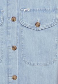 Jasnoniebieska koszula dżinsowa z dwiema kieszeniami na piersi, brązowymi guzikami i metką z logo Lee. Materiał ma miękką fakturę z widocznym przeszyciem.