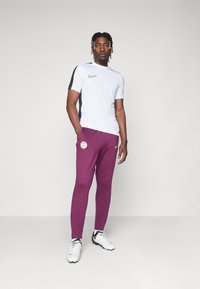 Nike rövid ujjú fehér sportpóló fekete akcentusokkal és lila melegítőnadrággal, amelyen logó található. Fehér és fekete stoplis cipővel viselve.