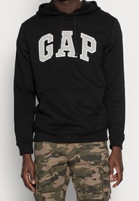 Sweat à capuche noir avec lettrage gris "GAP", poche kangourou et capuche à cordon. Porté avec un pantalon cargo camouflage. Matière en mélange de coton.