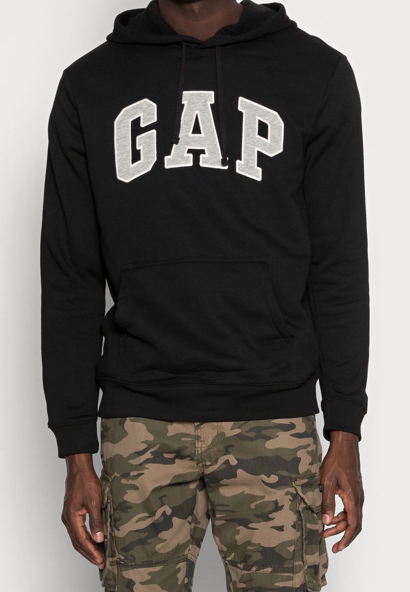 Sweat à capuche noir avec lettrage gris "GAP", poche kangourou et capuche à cordon. Porté avec un pantalon cargo camouflage. Matière en mélange de coton.