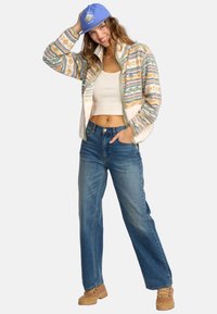 Chaqueta de forro polar con cremallera en tonos pastel y patrones abstractos, combinada con jeans de denim azul de tiro alto y botas marrones. Gorra azul en la cabeza.