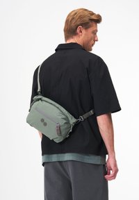 Grüne gepolsterte Schultertasche mit zwei runden Akzenten, verstellbarem Tragegurt, vorderer Reißverschlusstasche und einer glatten Textur. Auf dem Rücken getragen.