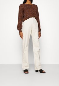 Brun långärmad blus med blå blommönster, i kombination med högmidjade krämfärgade byxor och svarta öppna blockklacksandaler.