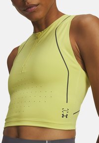Žltý športový crop top s predným zipsom, bodkovanou textúrou pozdĺž tela a kontrastnými čiernymi švami. Na spodnej pravej strane sa nachádza logo.