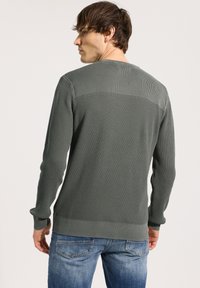 Pull gris en maille côtelée avec manches longues et col rond, présentant une finition texturée et un panneau arrière lisse contrastant. Associé à un jean bleu.