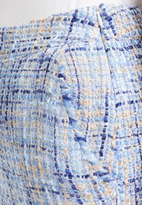 Gros plan sur un tissu tissé texturé aux tons bleu, beige et blanc, montrant une couture de poche avec un détail de bouton subtil.