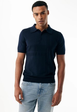 Hombre con una camiseta tipo polo de manga corta azul marino y jeans azul claro de pie contra un fondo blanco.