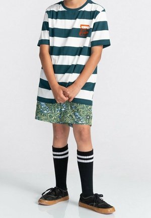 Enfant portant un t-shirt rayé vert et blanc, un short vert à motifs, des chaussettes noires montantes à rayures blanches et des baskets noires.