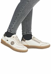 Bílé tenisky s hnědými gumovými podrážkami, texturovanými panely, kovovými akcenty a dekorativním logem. Navrženy pro pohodlí a každodenní nošení.