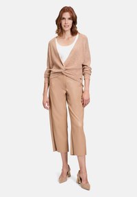 Beige Strickcardigan mit verdrehtem Vorderteil, über einem weißen Oberteil getragen; kombiniert mit hellbraunen Leder-Culottes und beigen Schuhen mit goldenen Akzenten.