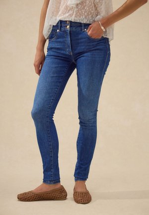 Žena oblečená v modrých úzkych džínsoch, bielom čipkovanom topu, hnedých pletených balerínkach a striebornom náramku, stojaca s jednou rukou v vrecku.