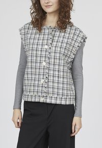 Ternet, ærmeløs vest i sort og hvid, med frynser og store knapper, båret over en langærmet grå top.