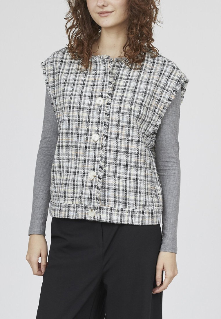 Ternet, ærmeløs vest i sort og hvid, med frynser og store knapper, båret over en langærmet grå top.