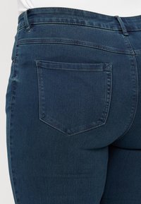 Dark blue denim jeans with a mid-rise waist, featuring a single back pocket. Smooth texture with subtle stitching details.