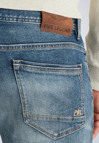 Jeans de mezclilla con un lavado azul desgastado, con un parche de cuero marrón de la marca que lleva grabado "PME LEGEND" y costuras detalladas en los bolsillos.