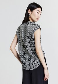 Giovane donna con capelli neri lisci che indossa un top senza maniche con motivo geometrico in bianco e nero, pantaloni neri e orecchini a goccia in argento.