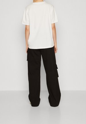 Persona que lleva una camiseta blanca lisa de manga corta y pantalones cargo negros y holgados, de pie frente a un fondo claro y sencillo.