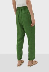 Woman wearing green patterned pants with elastic waist and beige sandals, standing against a plain gray background.