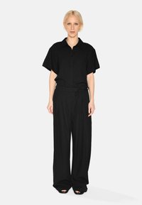 Chemise noire à manches courtes avec un bouton sur le devant et une poche, assortie à un pantalon noir à larges jambes, avec taille nouée et tissu lisse.