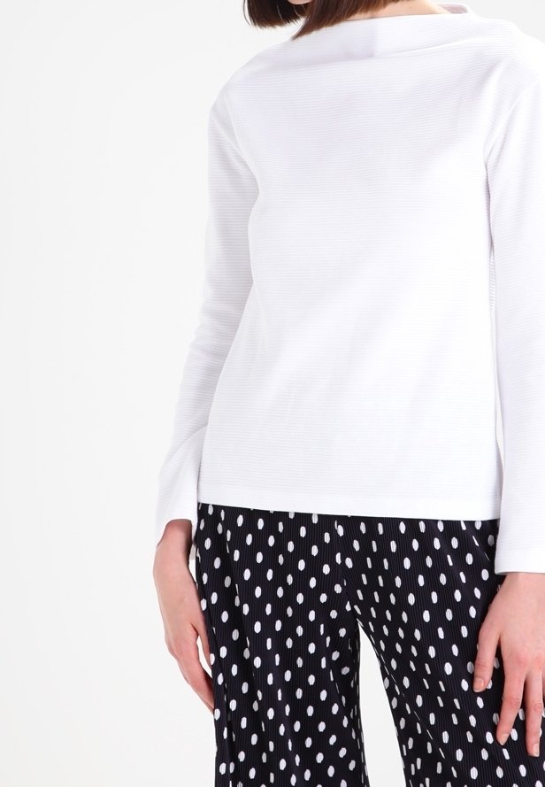 Haut blanc à manches longues avec une texture côtelée et un décolleté bateau, associé à un pantalon noir et blanc à pois avec une coupe droite.