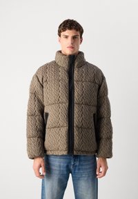 Polstret pufferjakke i brun med kabelstrik tekstur, høj krave, lynlås lukning foran og sidelommer, båret over blå denim jeans.