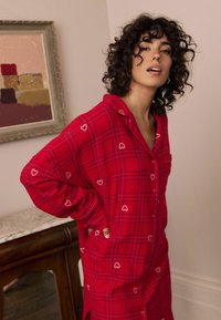 Czerwona koszula nocna w kratę z białymi wzorami serc, z guzikami z przodu, kołnierzykiem i długimi rękawami. Miękka, szczotkowana tkanina; luźny krój.