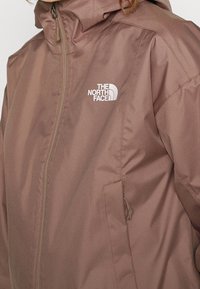Bruine waterdichte jas met een capuchon. Voorzien van een ritssluiting aan de voorkant en zijzakken. Het logo van The North Face is in het wit op de borst weergegeven.