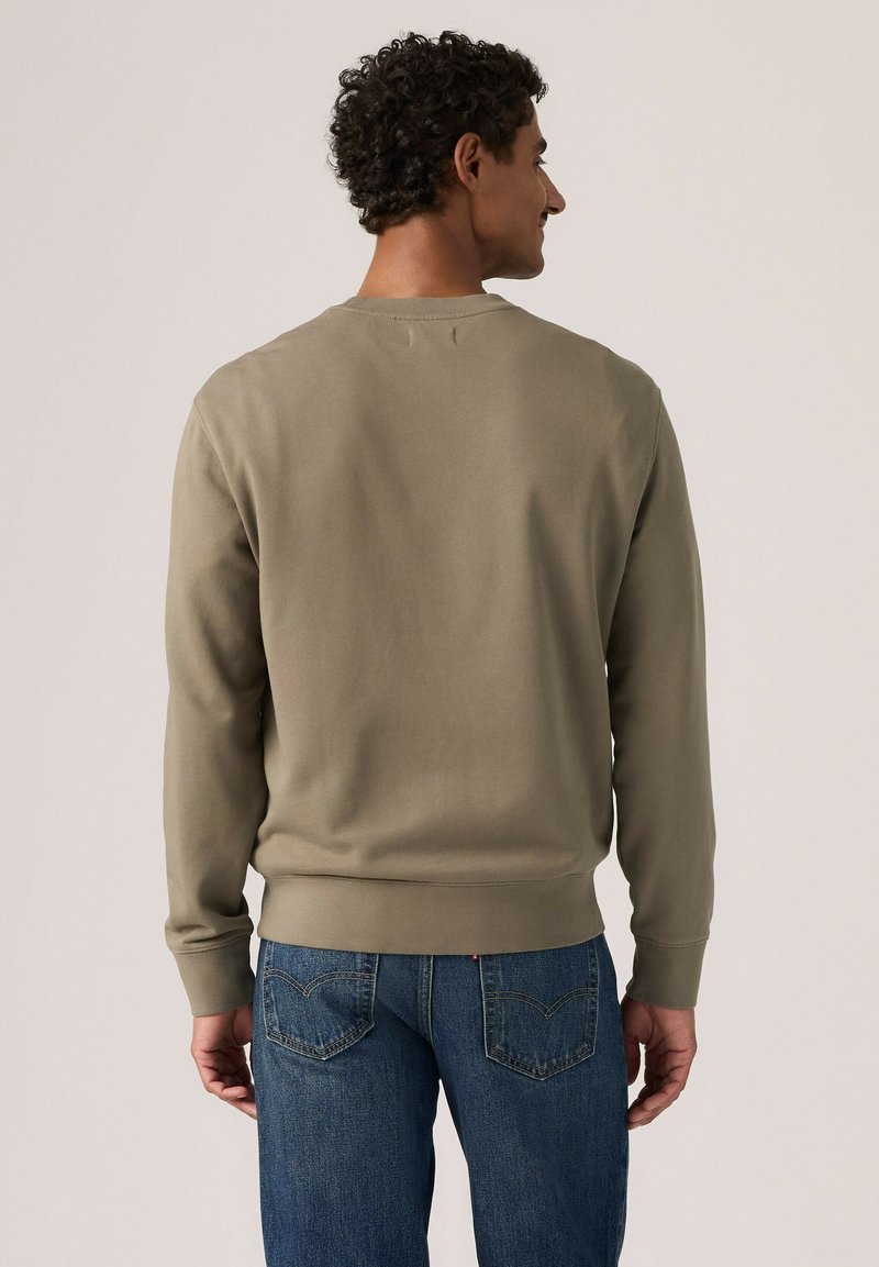 Mann mit lockigem Haar, der einen schlichten olivgrünen Pullover und blaue Jeans trägt, mit dem Rücken zu einem neutralen Hintergrund.
