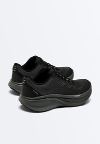 Para czarnych butów sportowych z grubymi szarymi podeszwami, dzianinowymi cholewkami i sznurowadłami, ustawionych obok siebie na białym tle.