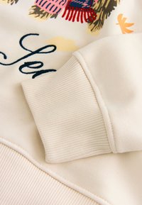 Manche de sweat-shirt couleur crème avec bord-côte côtelé, motif partiel floral et texte en bleu, rose, rouge, jaune et noir sur le tissu.