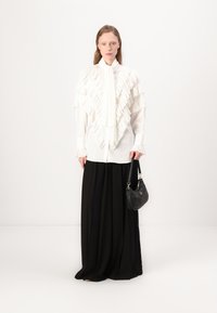 Blusa blanca con volantes en capas, combinada con una falda negra y fluida. La persona sostiene un pequeño bolso negro con una correa. Fondo simple.