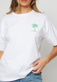 Mujer vestida con una camiseta blanca que tiene un pequeño gráfico de una palmera verde en el lado izquierdo del pecho, combinada con jeans de denim azules.