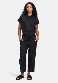 Chemise en lin noir à manches courtes et détail boutonné, assortie à un pantalon noir taille haute avec cordon de serrage et coupe décontractée. Sandales noires.