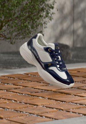 Biało-granatowy sneaker z grubą podeszwą, granatowymi sznurowadłami i złotym logo unoszący się nad podłogą o wzorze drewnianym i kamiennym.