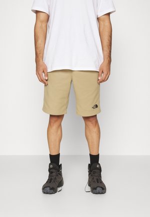 Man draagt beige The North Face shorts, wit T-shirt, zwarte sokken en zwarte wandelschoenen, staand tegen een witte achtergrond.