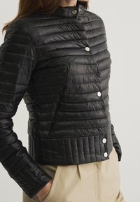 Svart puffjacka med kviltad design, ståkrage och tryckknappar. Har en figursydd silhuett och slät, blank yta.