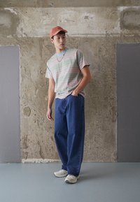 Camisa de manga corta a rayas en colores pastel, jeans azules, zapatillas beige y una gorra de color naranja claro, de pie contra una pared texturizada.