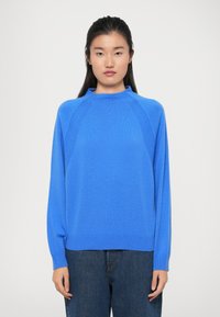 Blå strikkegenser med høy hals, raglanermer og ribbede mansjetter. Kombinert med mørkeblå denimjeans. Jevn tekstur.