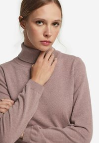 Jasnoróżowy sweter z golfem wykonany z miękkiej dzianiny, z dopasowanym krojem i ściągaczami na mankietach. Modelka delikatnie dotyka szyi.