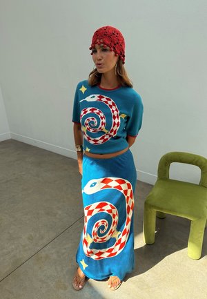 Naine, kes kannab sinist kootud topi ja seelikut punaste ja valgete spiraalse mustriga madu kujunditega, punast heegeldatud mütsi, seisab chartreuse'i värvi tooli kõrval minimalistlikus ruumis.