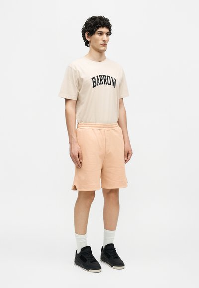 Άνδρας που στέκεται, φοράει μπεζ μπλούζα Barrow, ροδάκινο σορτς, άσπρες κάλτσες και μαύρα αθλητικά παπούτσια με λευκό φόντο.