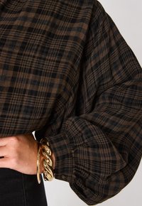 Camicia a quadri nera e marrone con maniche voluminose, polsini abbottonati e tessuto morbido, abbinata a un bracciale in oro.